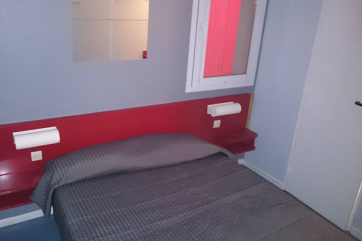 A small, narrow room with a single bed, a red headboard, and a window with a red curtain.