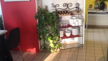 Una habitación con una pared roja, un cuadro enmarcado, un soporte metálico con tazas y platos, y un piso de baldosas.