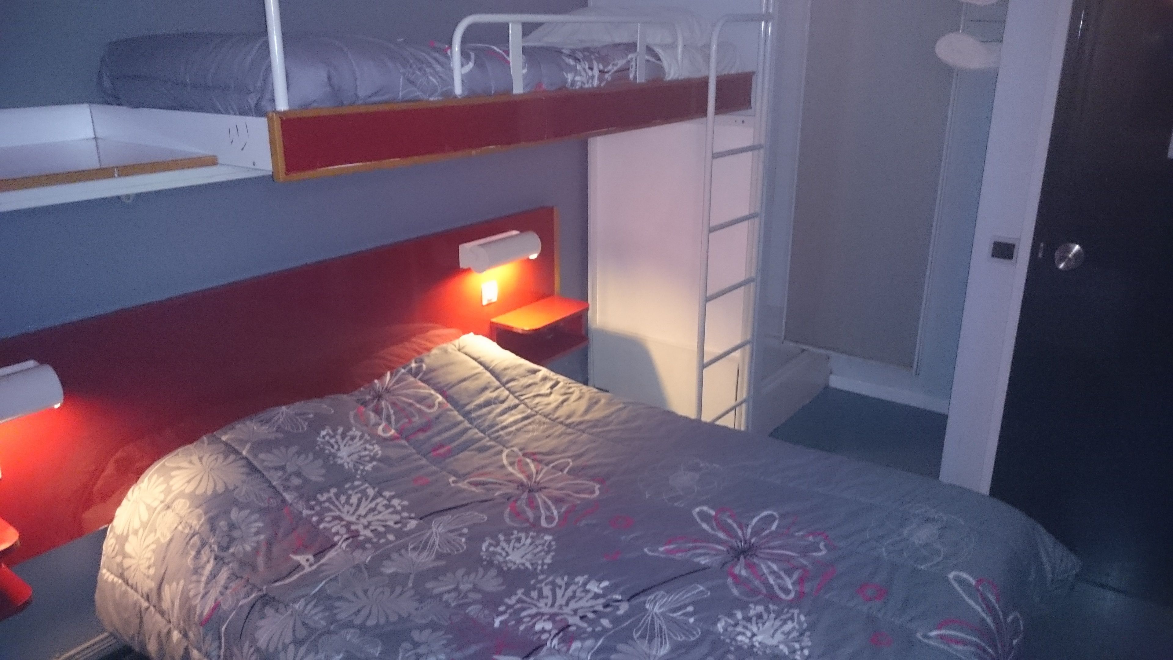 Una cama litera con una cama inferior y una cama individual, ambas con ropa de cama estampada. Hay una mesita de noche pequeña con una lámpara en la cama inferior.