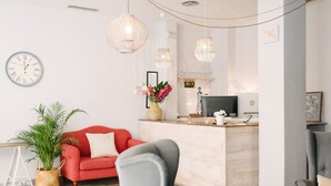 A modern living room with a red sofa, grey armchairs, a wooden reception desk, and a large clock on the wall. - The Boutike Guesthouse - Hostel (Seville)