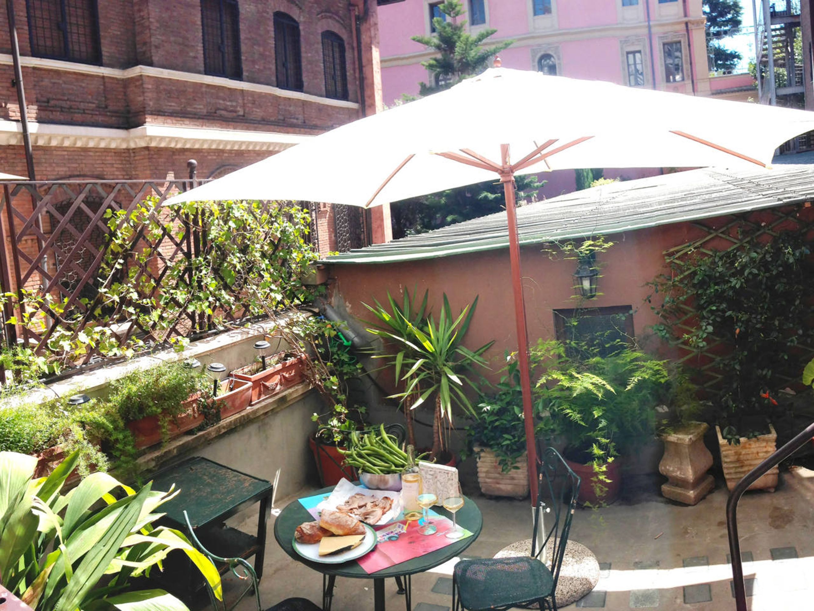 Una zona patio con un tavolo apparecchiato per due, circondata da piante in vaso e un ampio ombrellone.