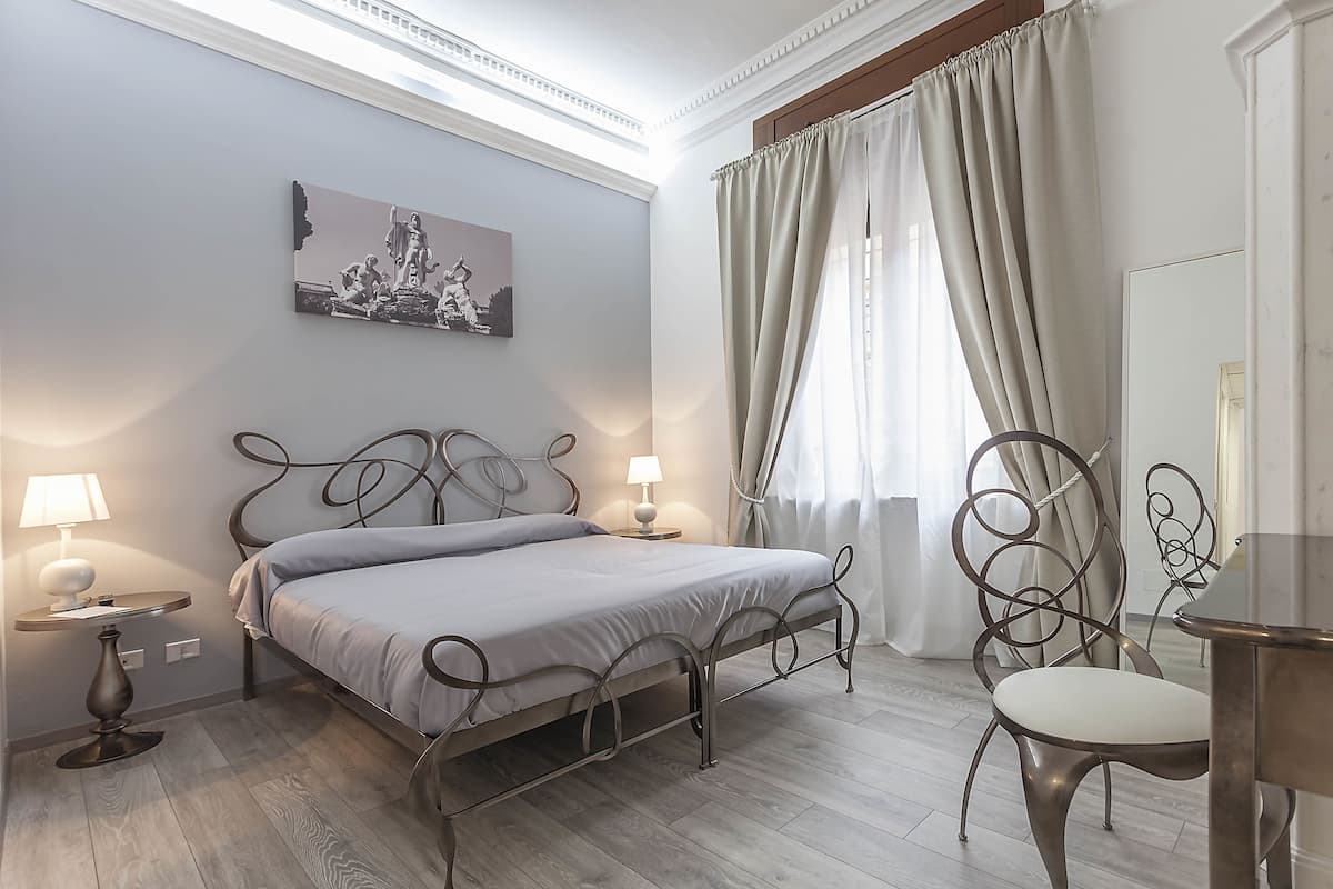 A bedroom with a bed, bedside lamps, a chair, a table, a painting, and a window with curtains.