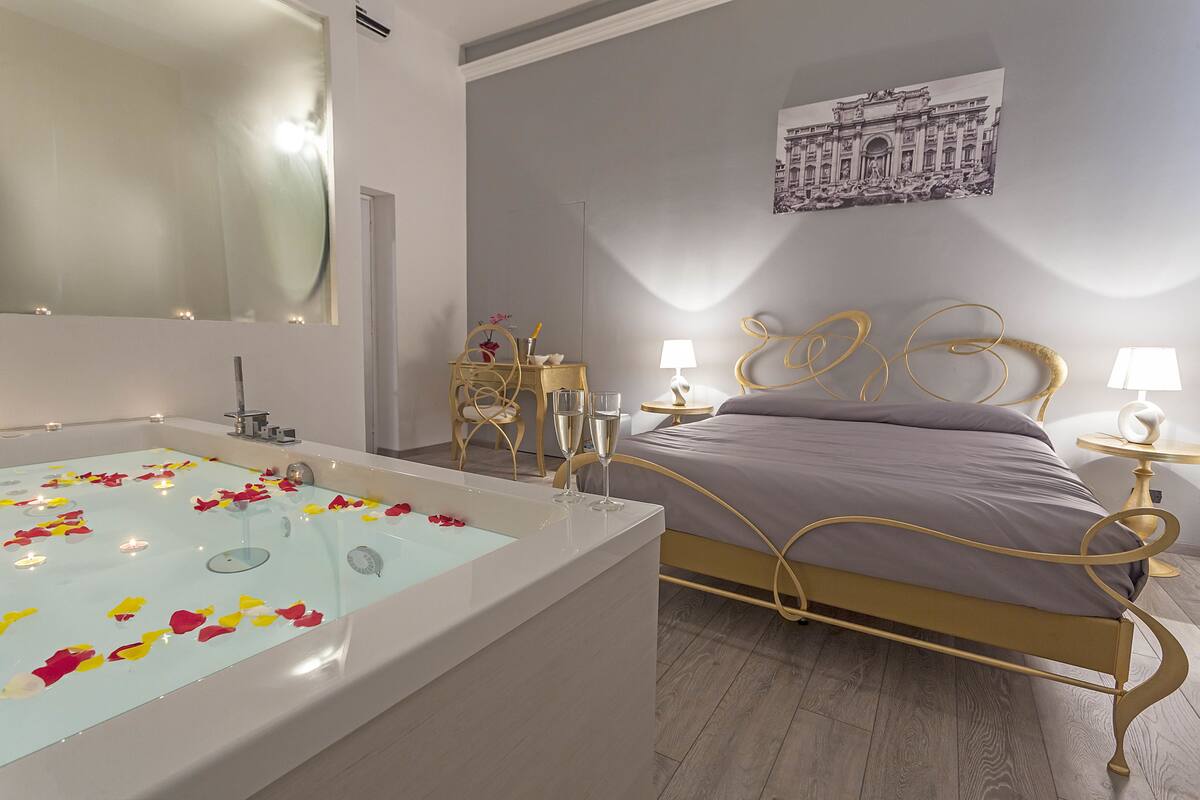 A modern bedroom with a large bathtub filled with rose petals, a bed with a gold frame, and a small bedside table with a lamp.