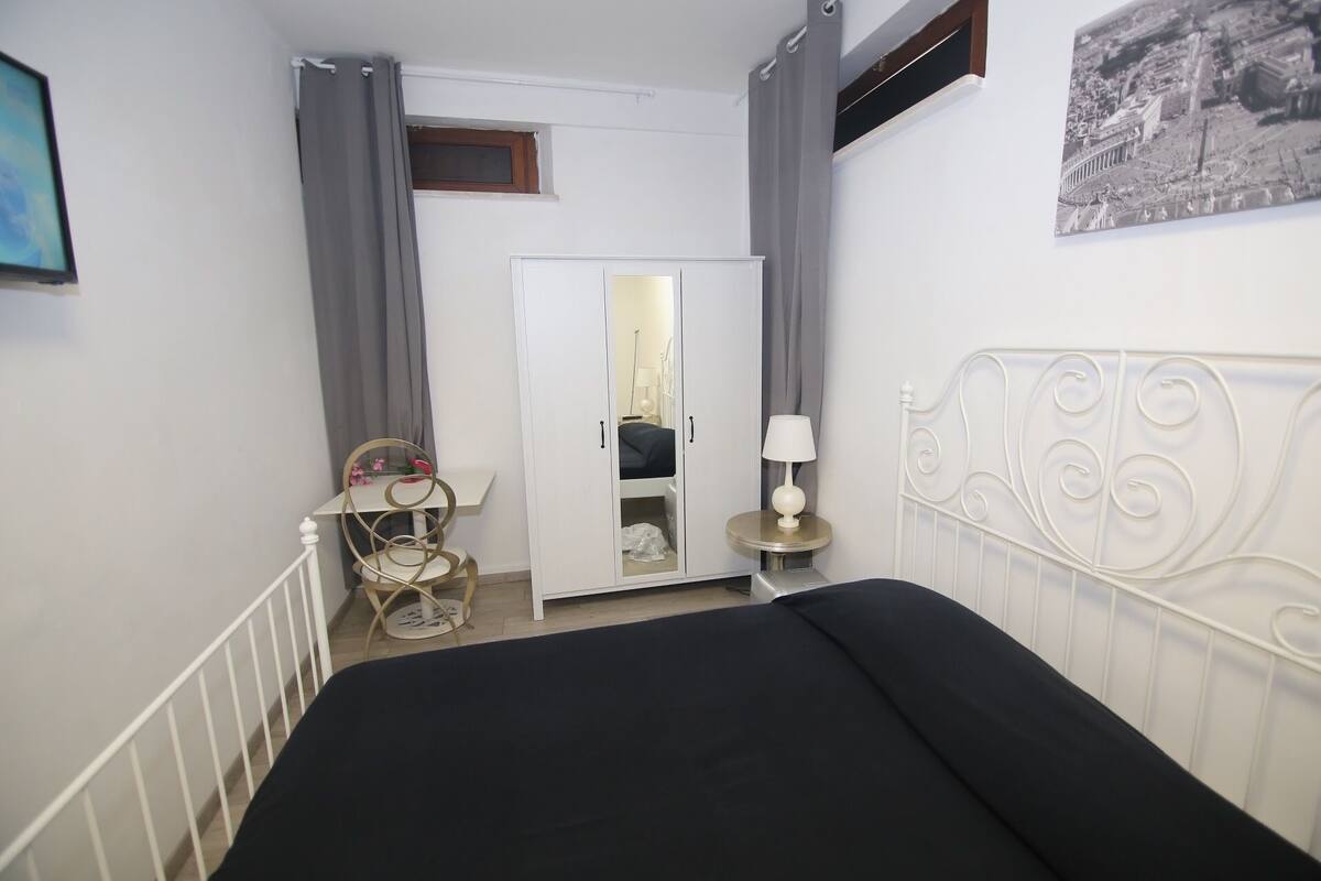 A bedroom with a bed, a bedside table with a lamp, a chair, a small table with a decorative item, and a door leading to another room.
