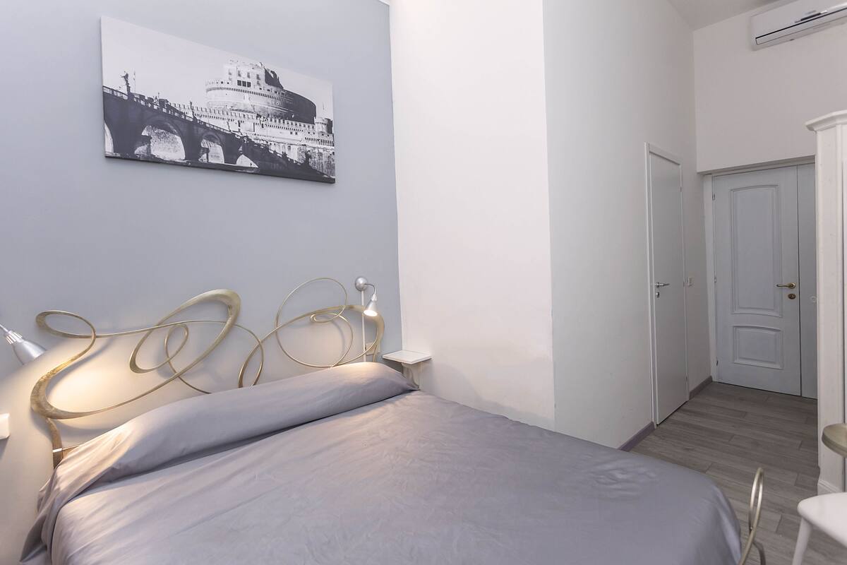 A bedroom with a bed, a bedside table with a lamp, a wall-mounted air conditioner, and a door leading to another room.