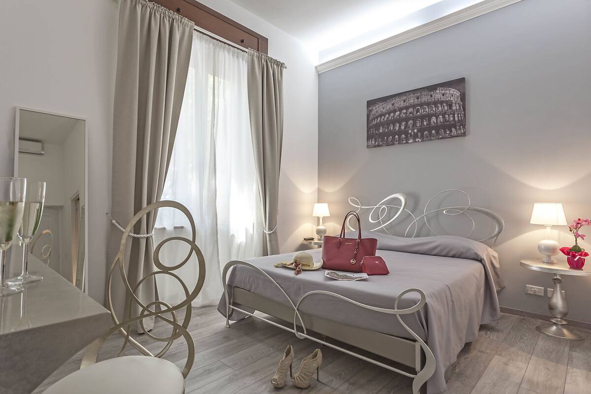 A bedroom with a bed, bedside lamps, a mirror, a curtain, a picture on the wall, and a bottle of champagne on a table.