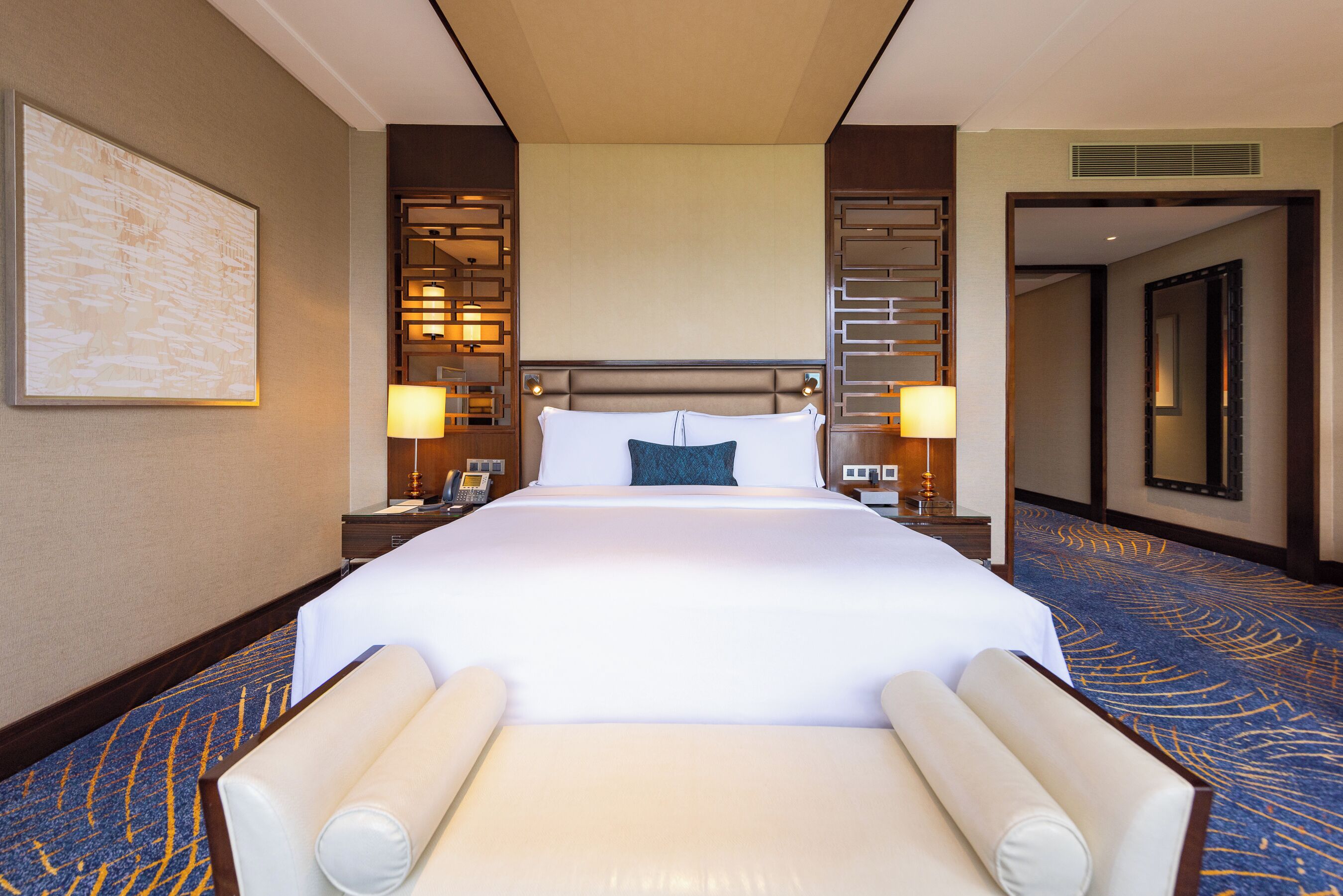 A hotel room with a large bed, two bedside lamps, a nightstand, a headboard with a built-in shelf, a large mirror, and a carpeted floor.