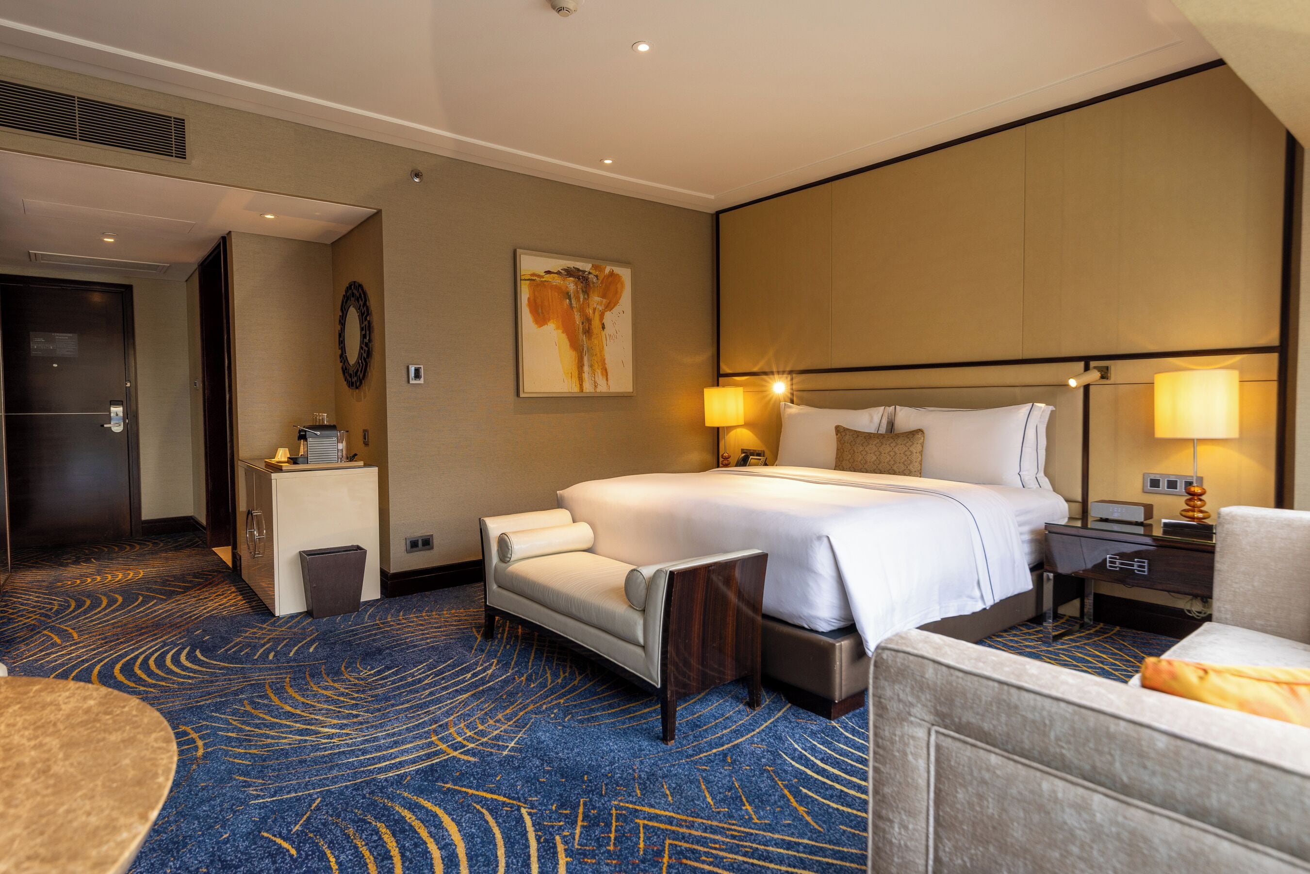Une chambre d’hôtel avec un grand lit, un bureau, une chaise, un petit meuble et un tapis à motifs géométriques.