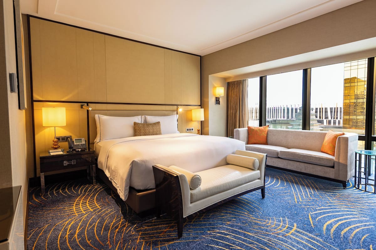 Habitación de hotel con una cama grande, un sofá, un escritorio con lámpara y vistas a la ciudad a través de amplias ventanas.