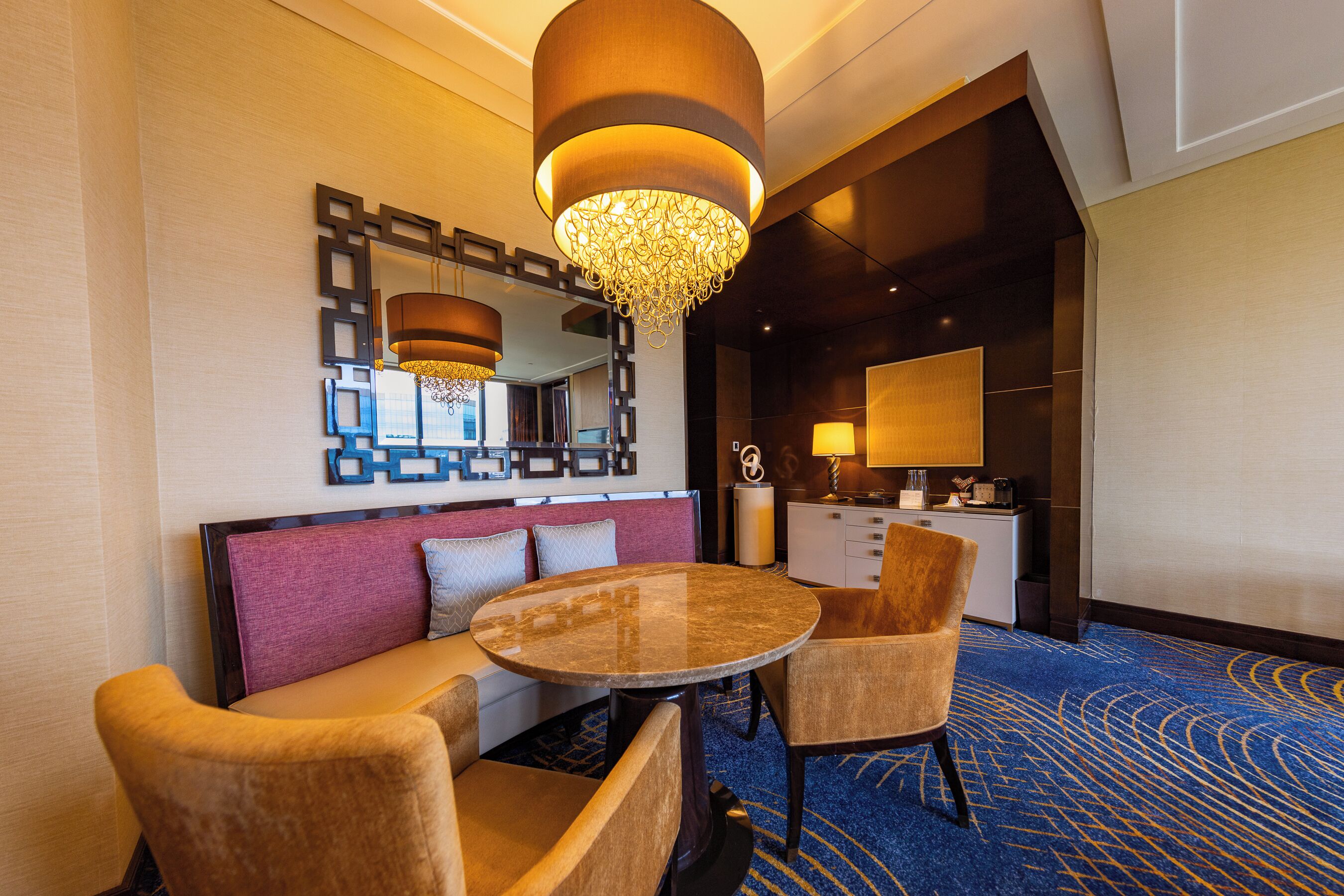 Un hall d’hôtel avec une table ronde, deux chaises, un canapé, un miroir décoratif et un lustre.