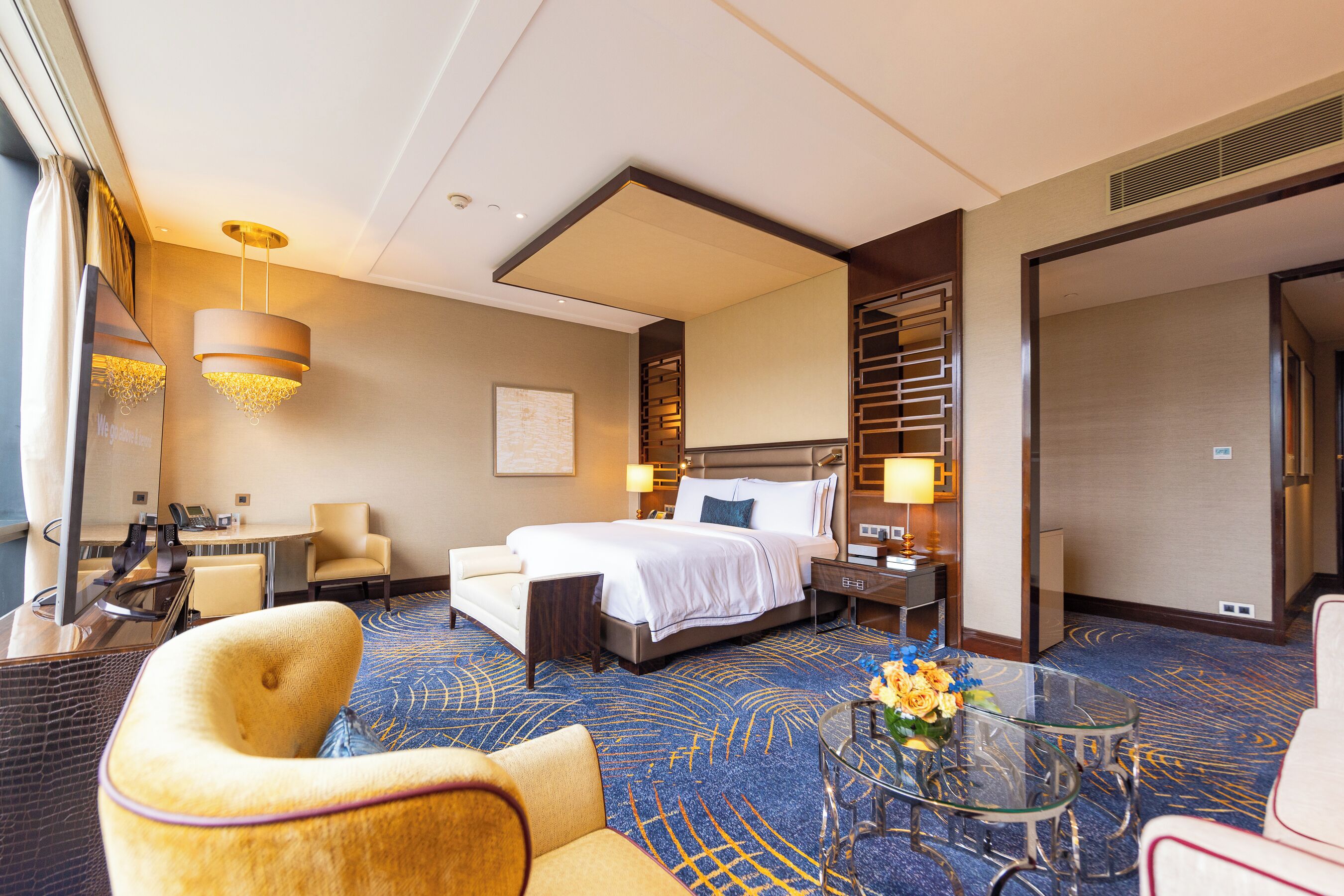 A hotel room with a bed, a desk, a chair, a glass coffee table, and a decorative vase.