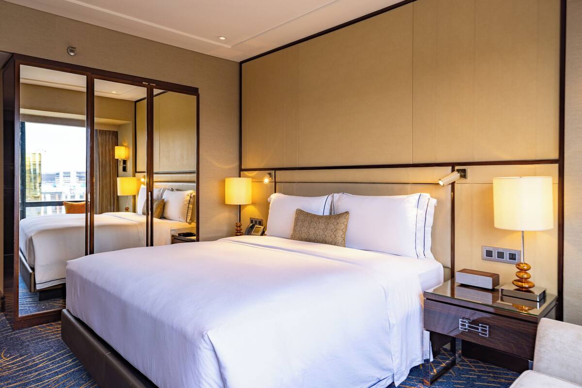Habitación de hotel con una cama grande, dos mesitas de noche con lámparas, un espejo y vistas a la ciudad.