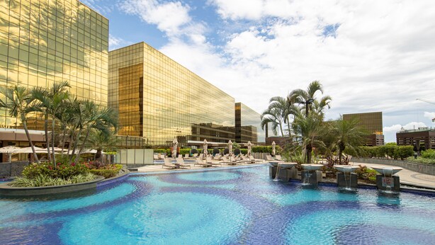 Una piscina con sillones de descanso y palmeras, rodeada de edificios modernos.