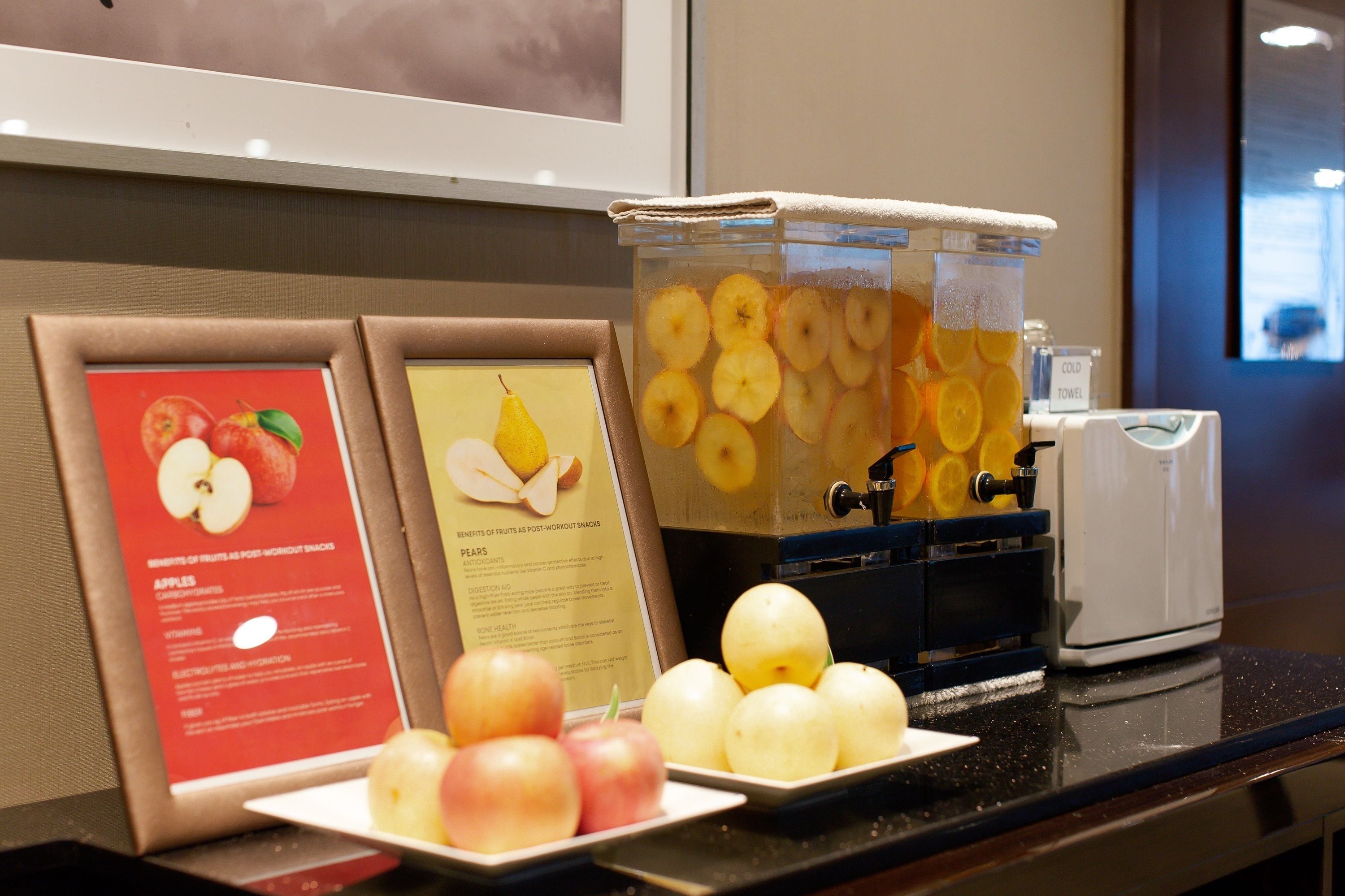A breakfast bar with a fruit display, including apples and oranges, and a juice dispenser.