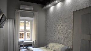 A bedroom with a bed, a desk, and a chair. There is an air conditioning unit on the wall and a window with curtains.