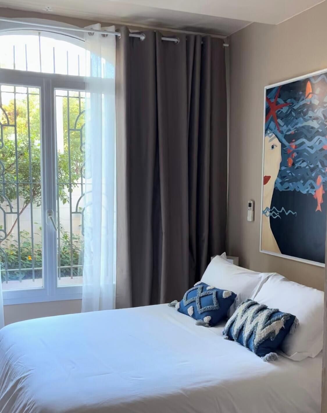 A hotel room with a bed, curtains, a window with a view of greenery, and a painting on the wall.