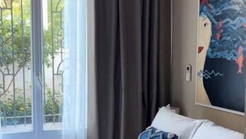 A hotel room with a bed, curtains, a window with a view of greenery, and a painting on the wall.