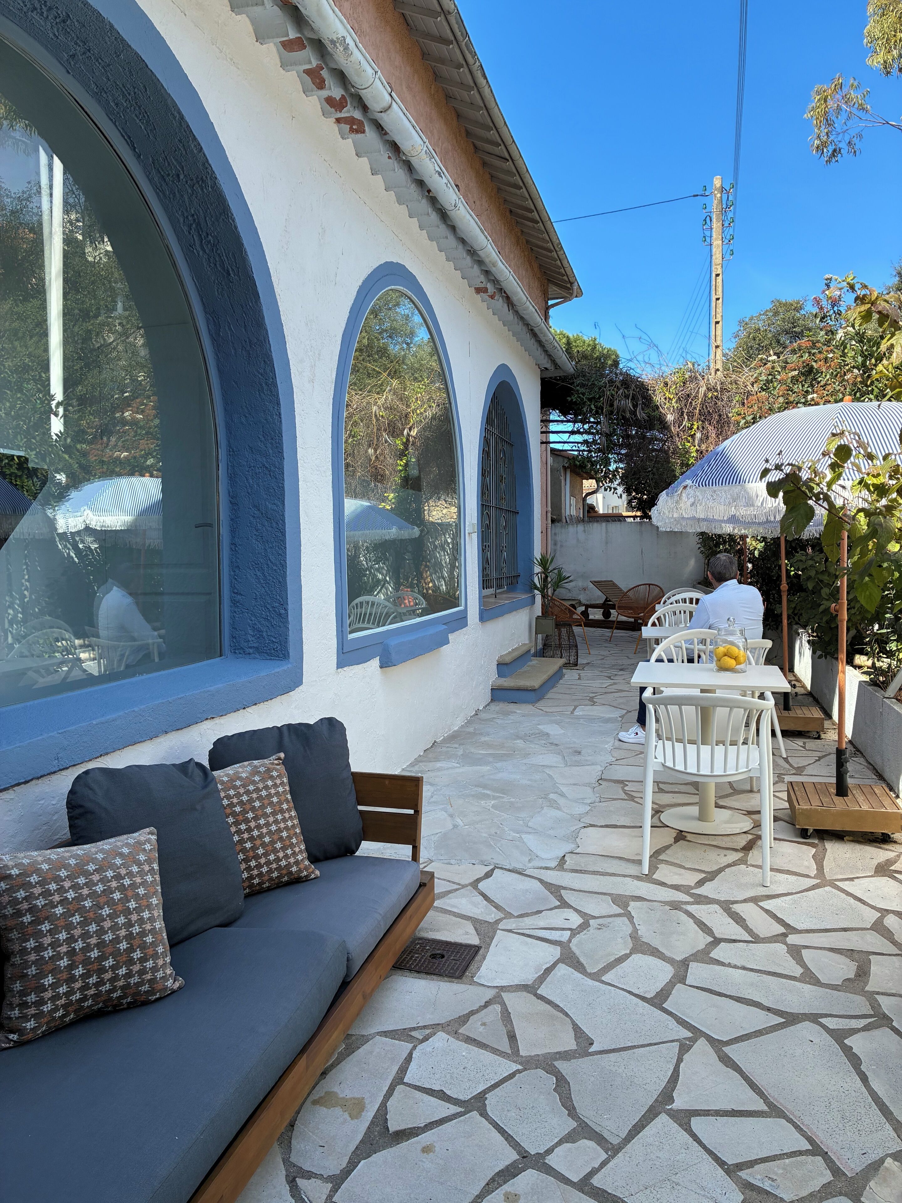 Eine Terrasse mit Steinboden, einem weißen Tisch und Stühlen sowie einem großen Fenster mit blauer Umrandung.