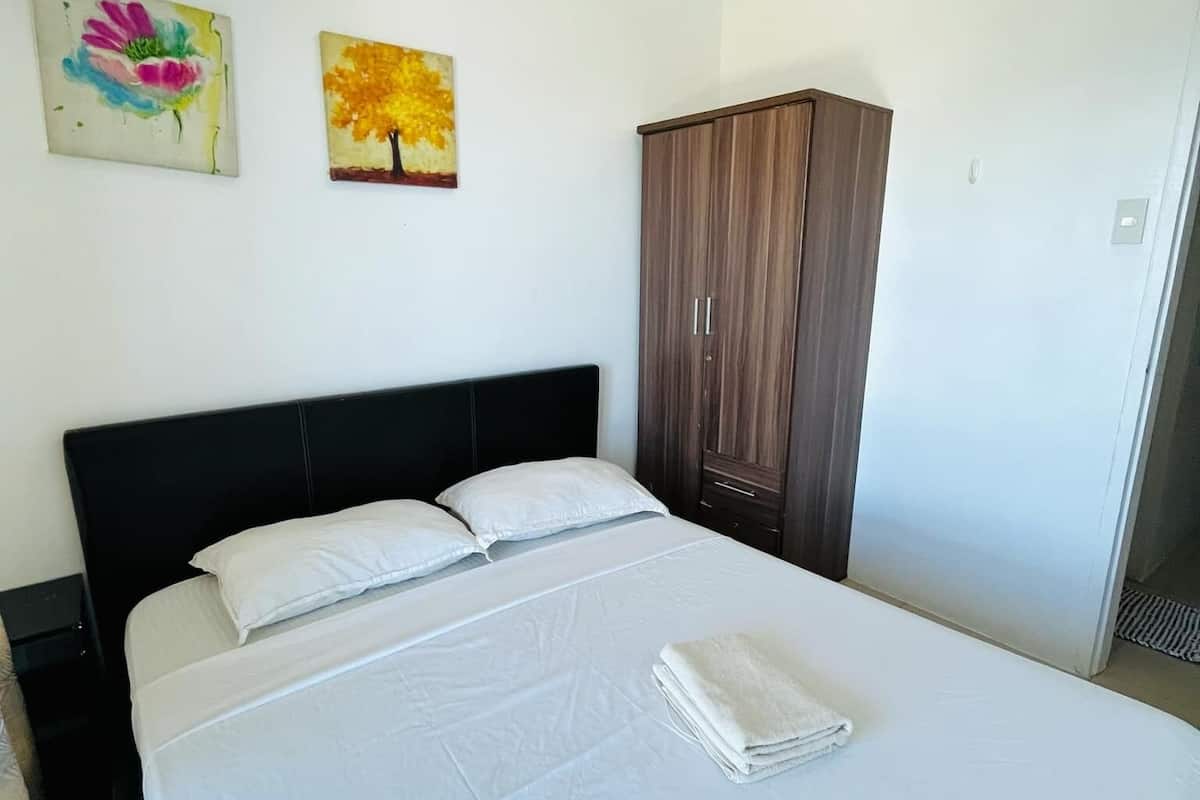 A neatly made bed with white linens and a folded towel, a wooden wardrobe, and two wall paintings.
