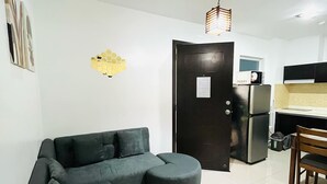 A modern living room with a sectional sofa, a small ottoman, a dining table with chairs, a kitchen area with appliances, and a decorative honeycomb wall art piece. - Primavera Residences (Cagayan de Oro)
