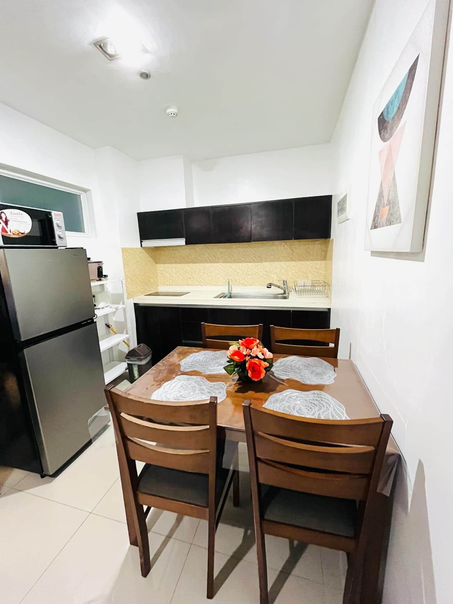 A compact kitchen with a refrigerator, microwave, and sink. There is a dining table with four chairs and a vase of flowers. The kitchen has dark cabinets and white walls.