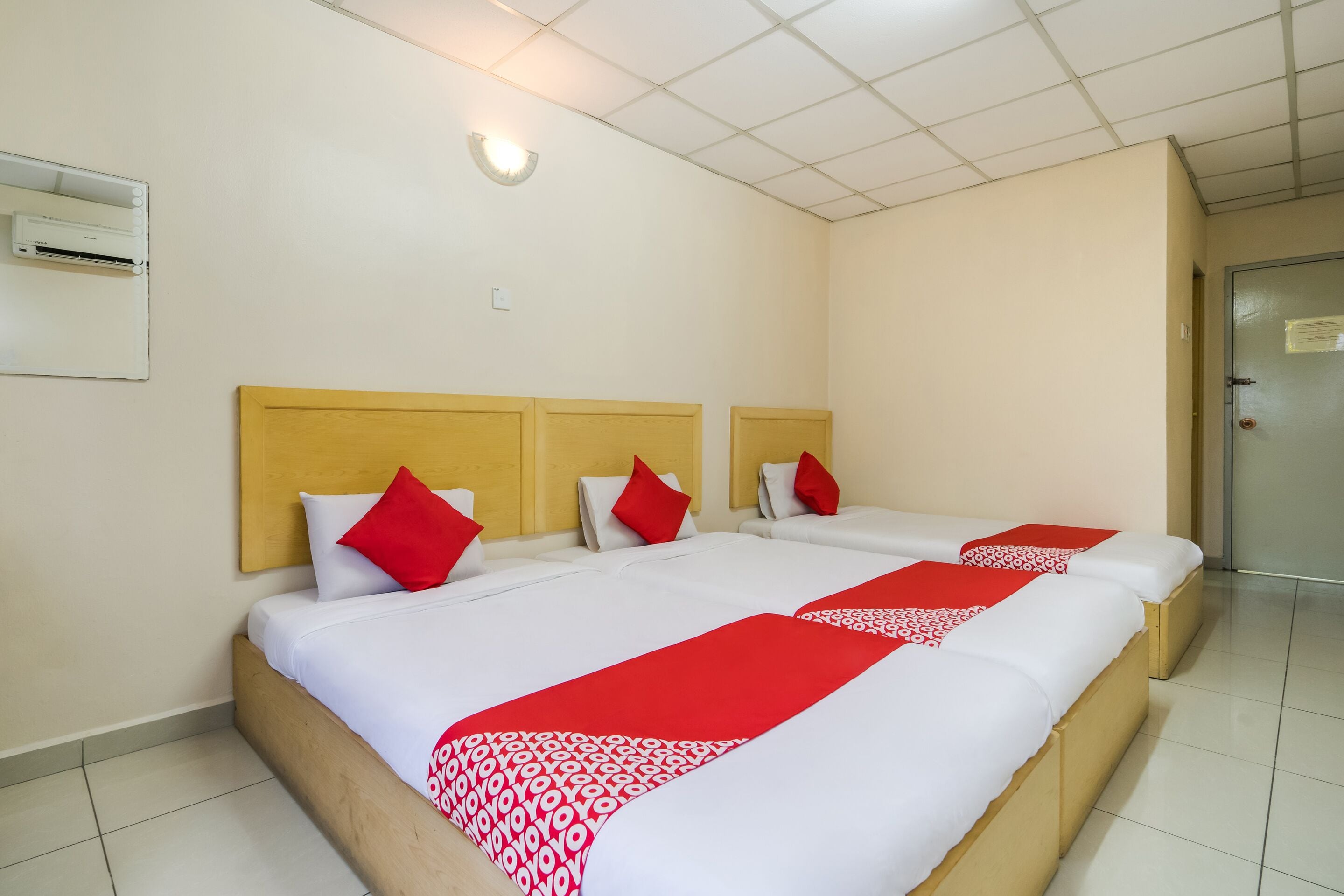 Une chambre d’hôtel avec trois lits, chacun doté de draps blancs et d’éléments décoratifs rouges, d’une tête de lit en bois et d’un sol carrelé.