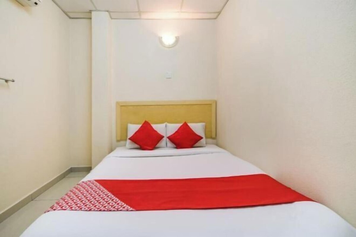 A narrow hotel room with a single bed, a wooden headboard, and a red throw blanket.