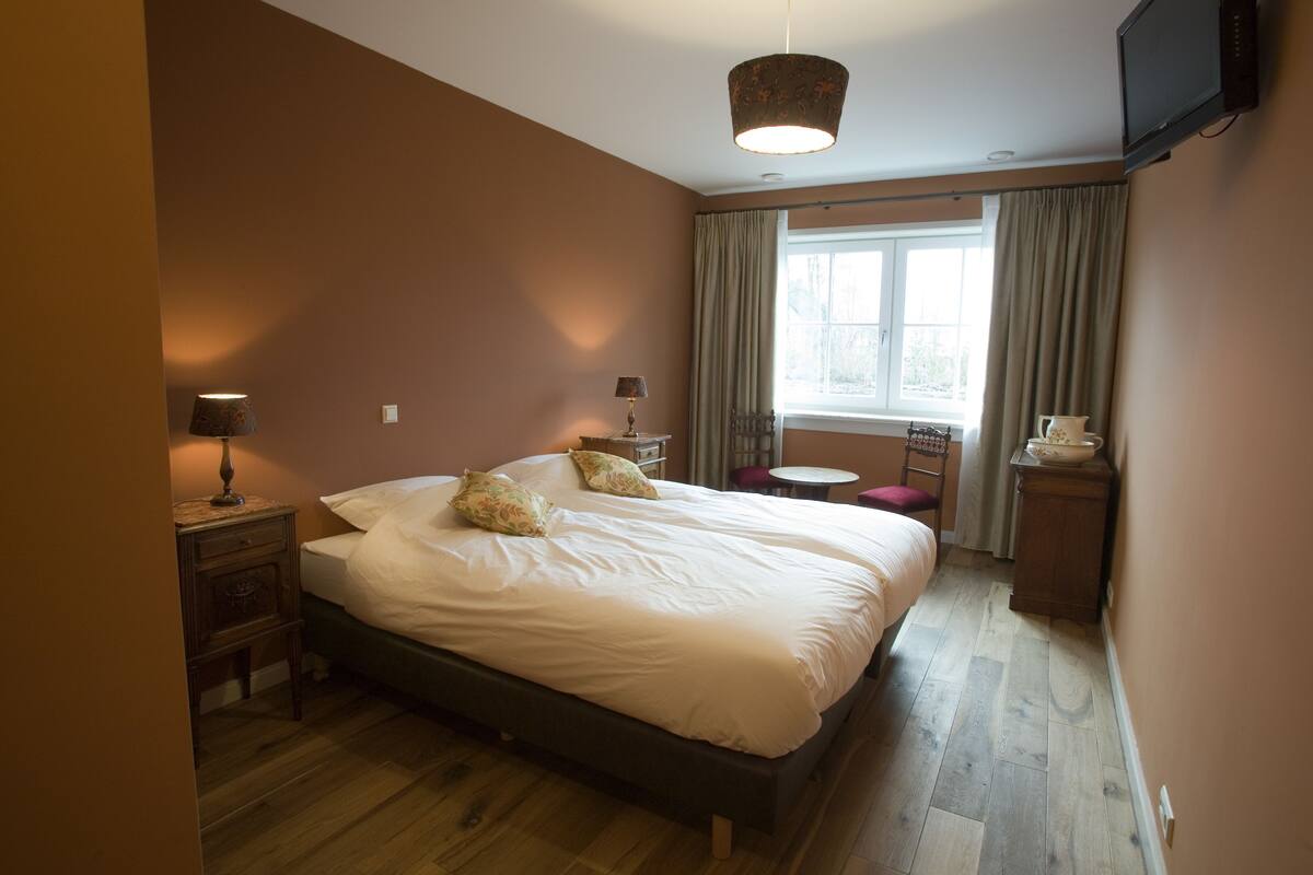 A bedroom with a large bed, bedside tables, a TV, a window with curtains, and a small table with a bowl.
