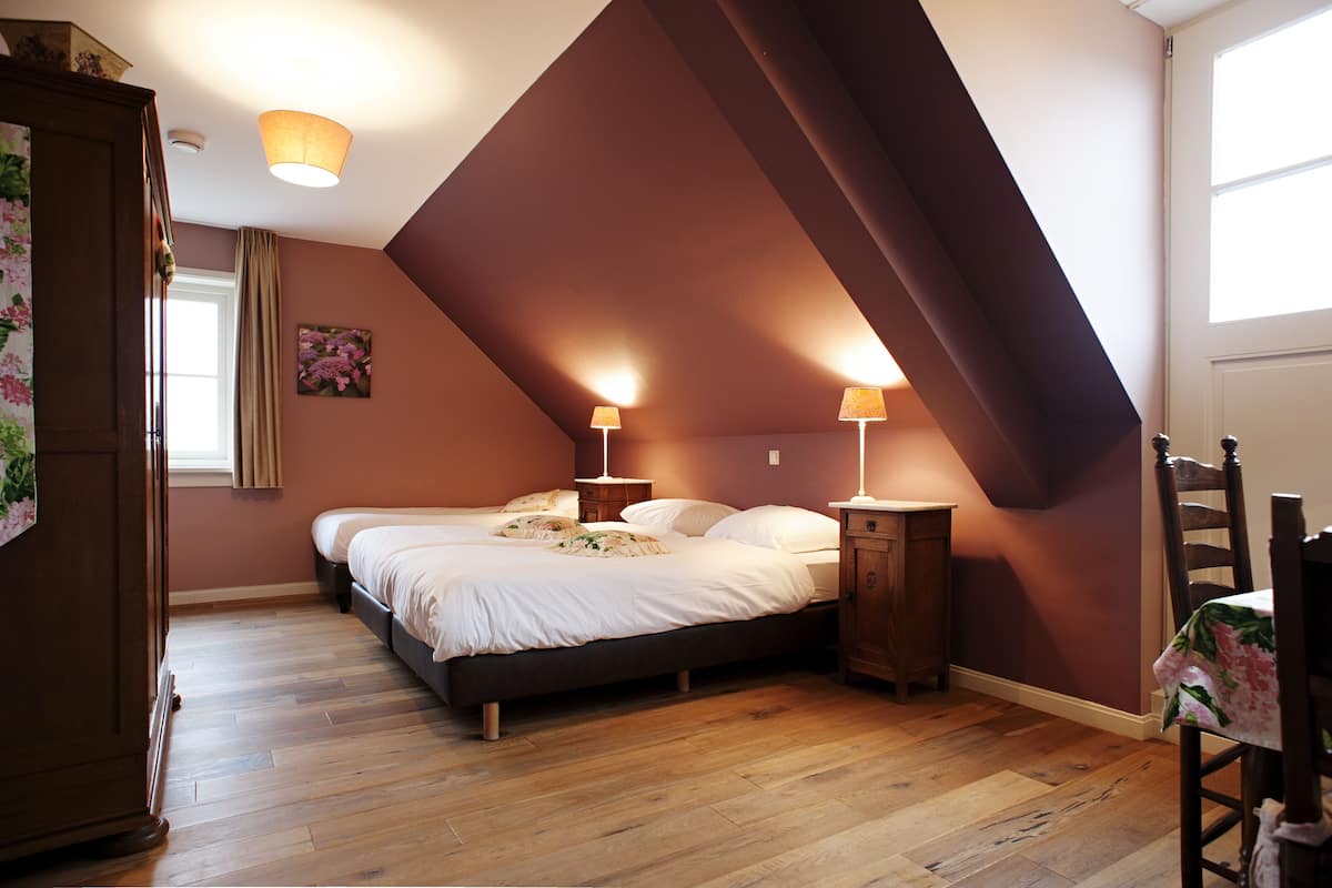 A bedroom with a sloped ceiling, a bed with white linens, two bedside lamps, a wooden wardrobe, and a window with curtains.