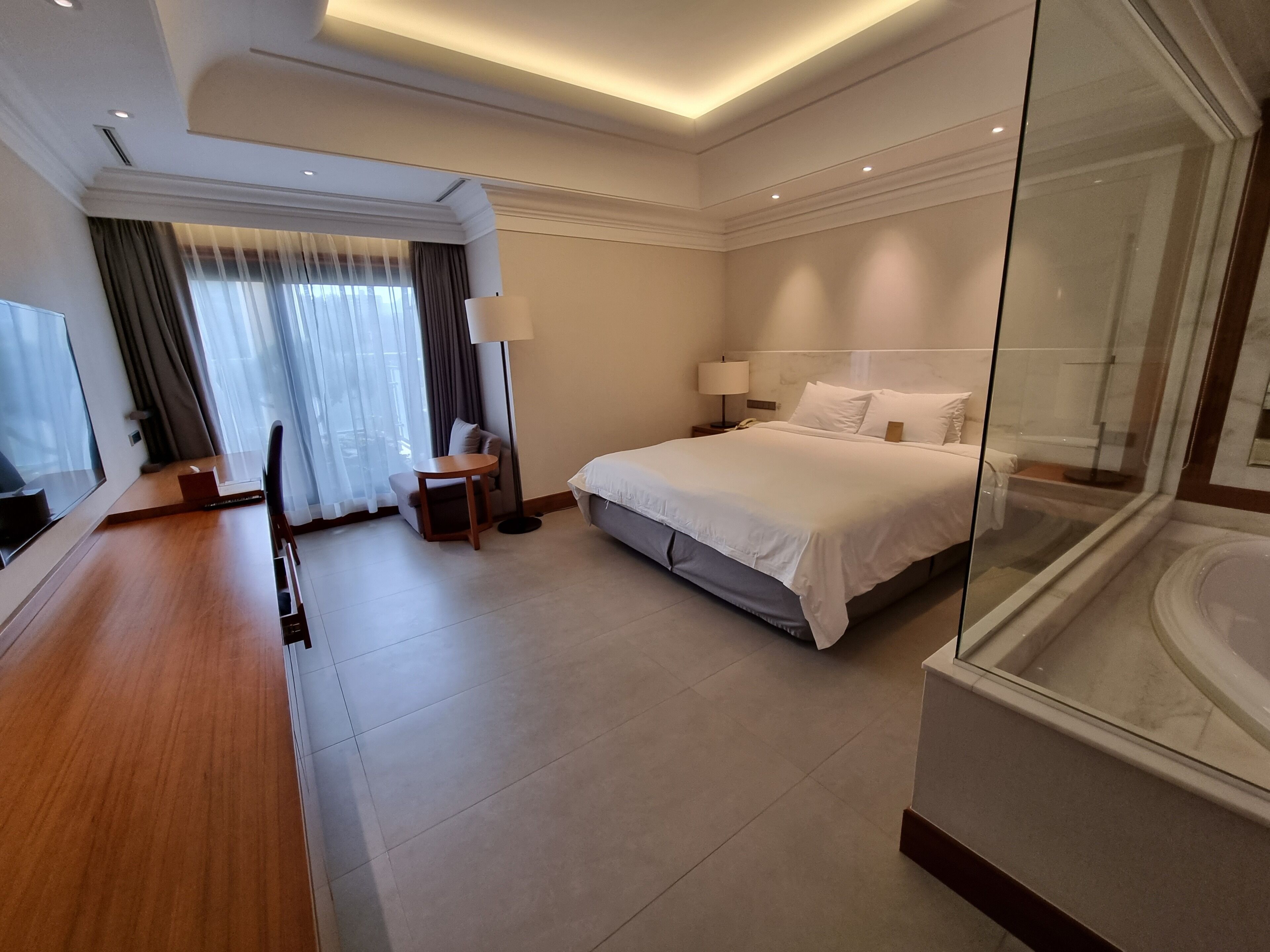 A modern hotel room with a large bed, a desk, a chair, a lamp, and a glass partition.
