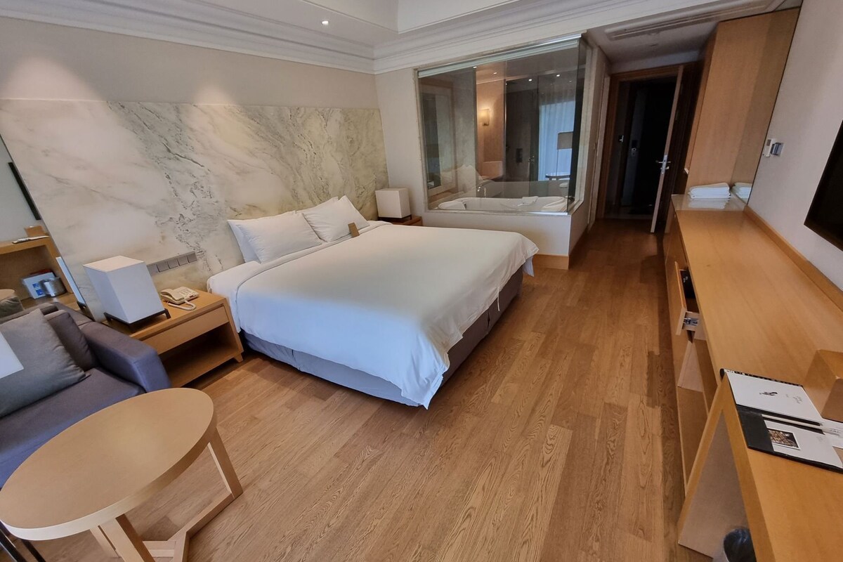 A hotel room with a large bed, a desk, a chair, a sofa, a TV, and a bathroom with a glass shower.