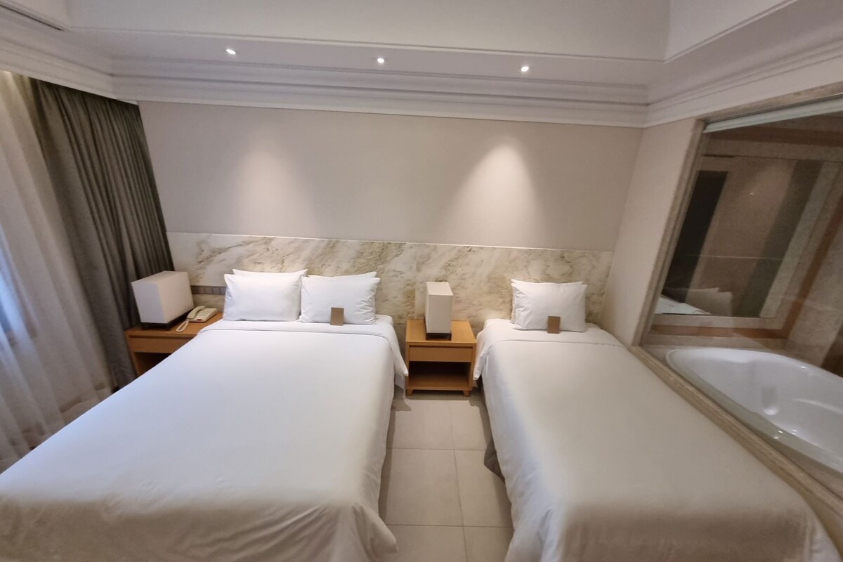 A hotel room with two single beds, a marble headboard, a bedside table with a lamp, a telephone, and a large window with a view.