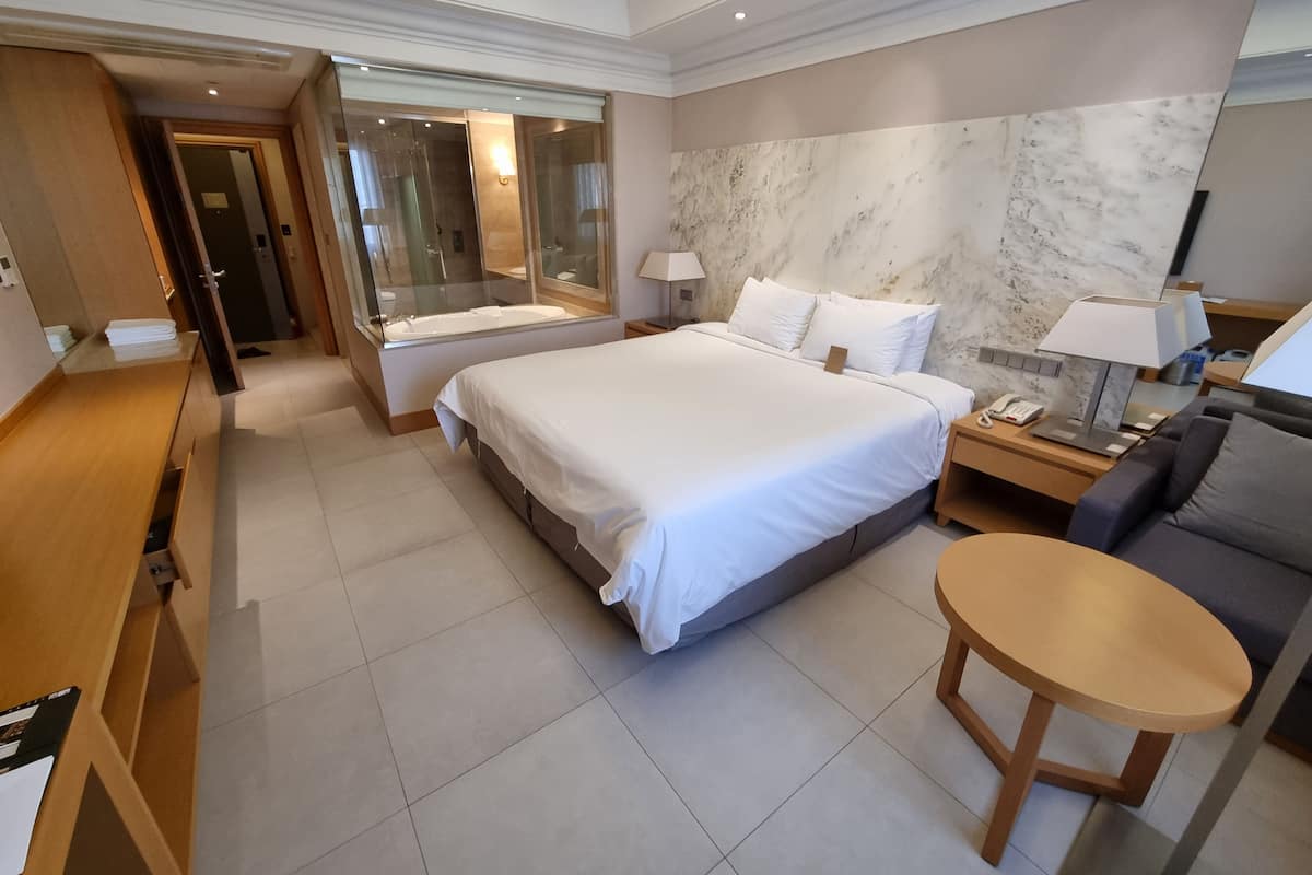 Una habitación de hotel moderna con una cama grande, un escritorio, una zona de estar y un baño visible a través de una puerta de cristal.
