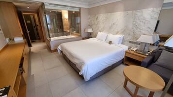 Una habitación de hotel moderna con una cama grande, un escritorio, una zona de estar y un baño visible a través de una puerta de cristal.
