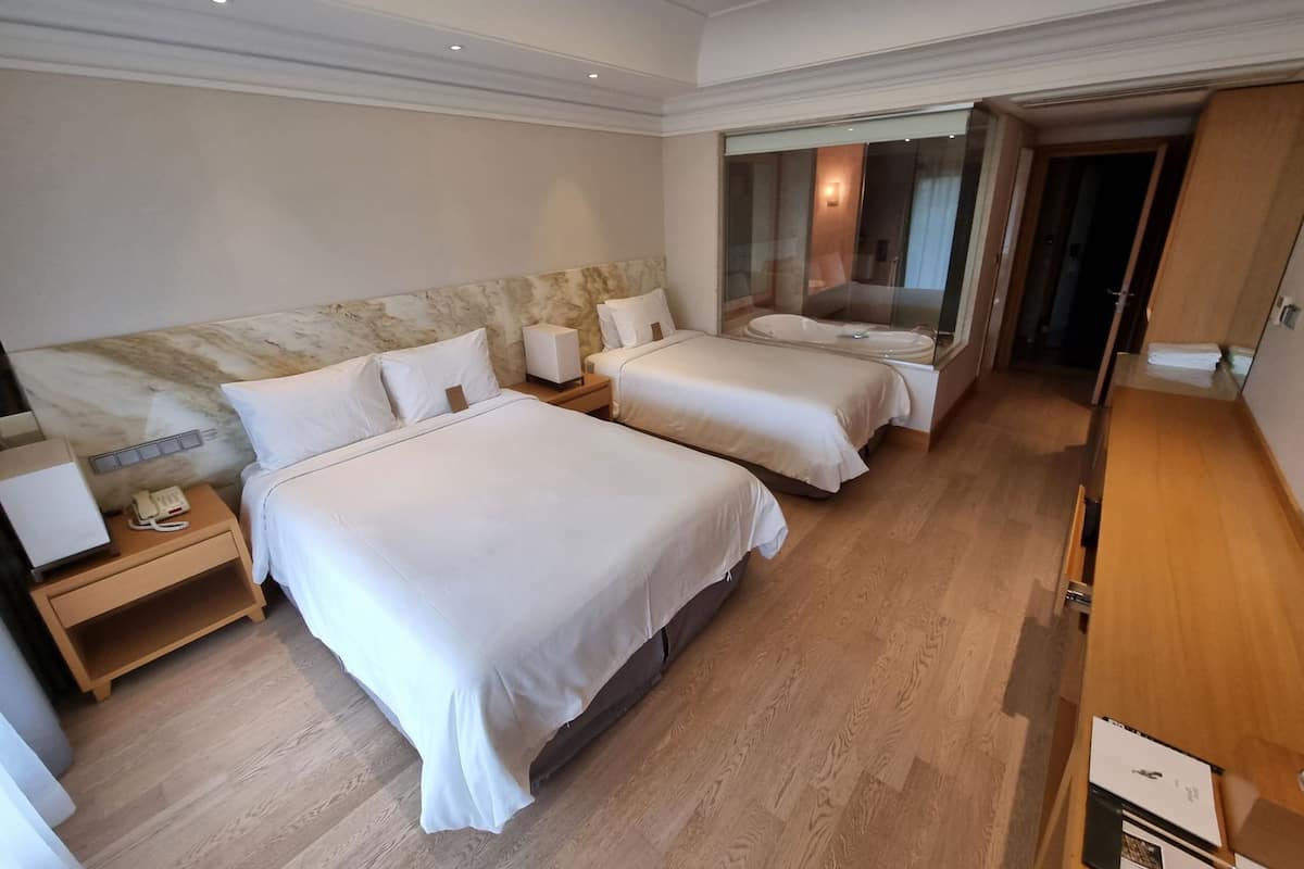Habitación de hotel con dos camas, cabecera de mármol, mesita de noche con teléfono y baño visible a través de una puerta de cristal.