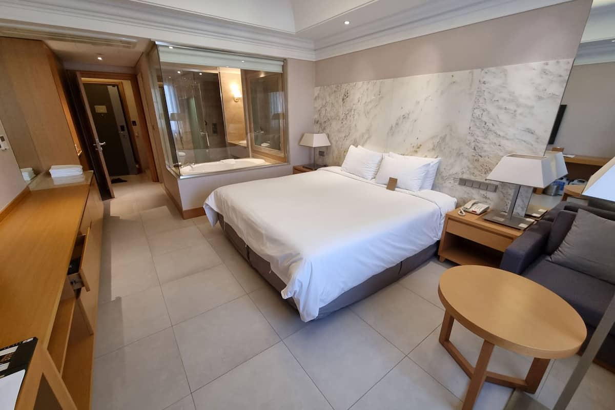 Una habitación de hotel moderna con una cama grande, un escritorio, una silla, una mesita y un baño con ducha de vidrio.