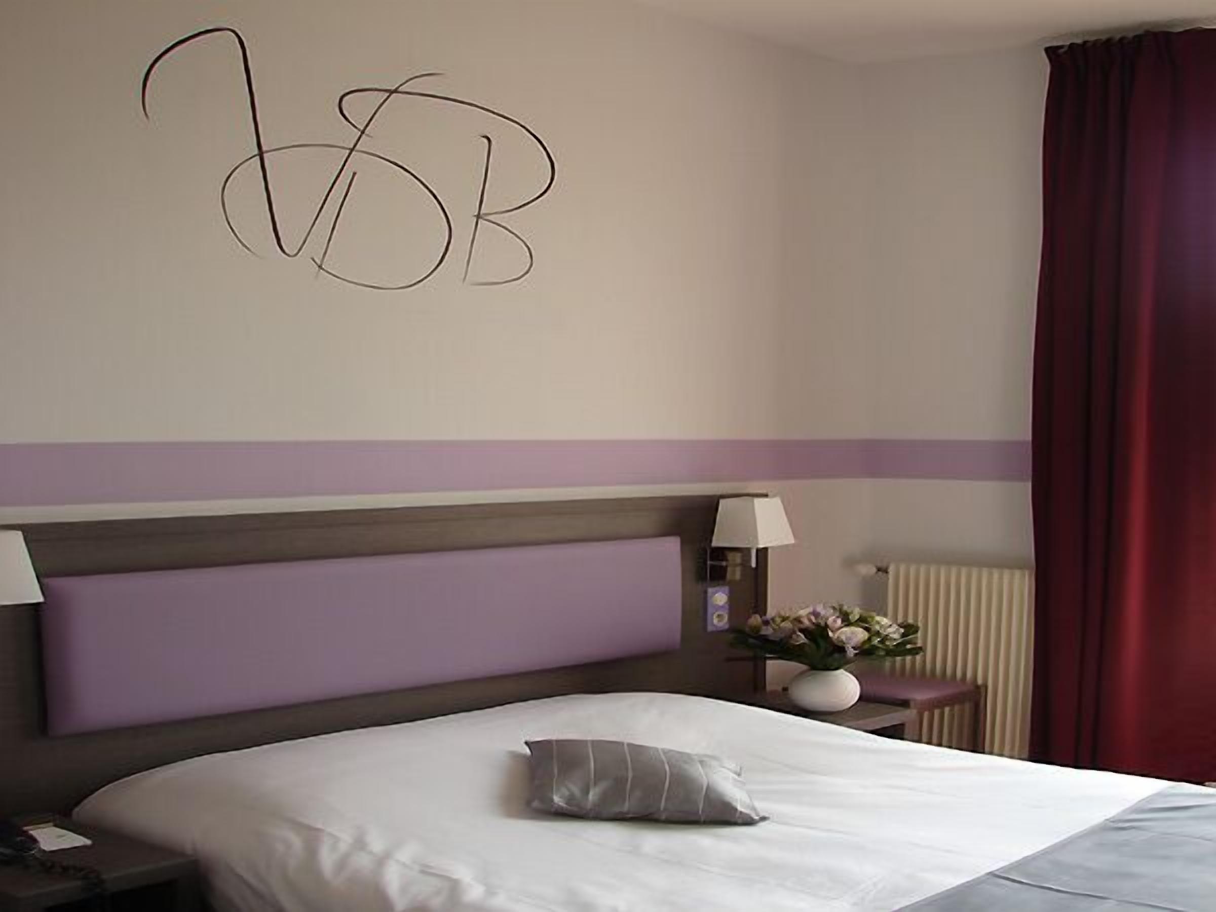 A hotel room with a large bed, a headboard, two bedside lamps, a nightstand, a vase with flowers, and a red curtain.