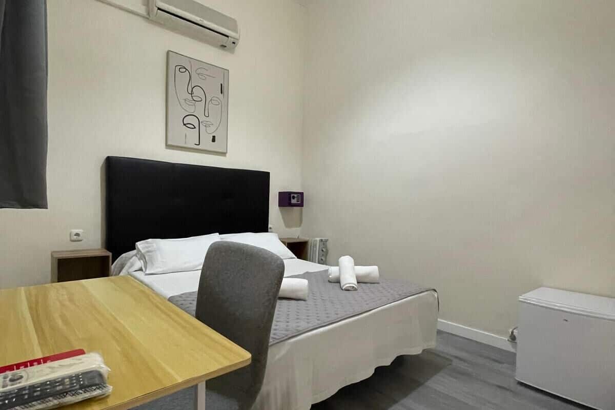 Una habitación de hotel con una cama, un escritorio, una silla, un equipo de aire acondicionado, una obra de arte en la pared y un radiador blanco.