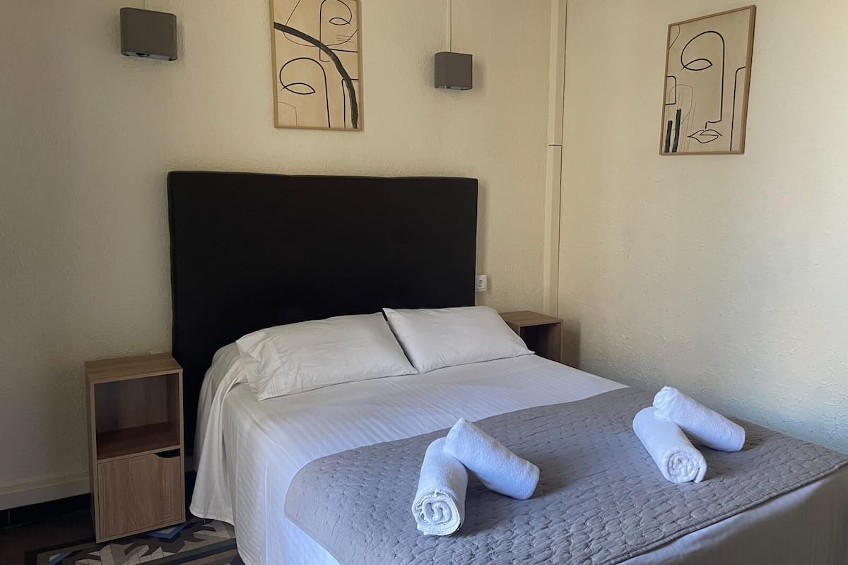 Una cama bien hecha con sábanas blancas y una colcha gris, dos mesitas de noche con detalles de madera y una obra de arte abstracto sobre la cama.