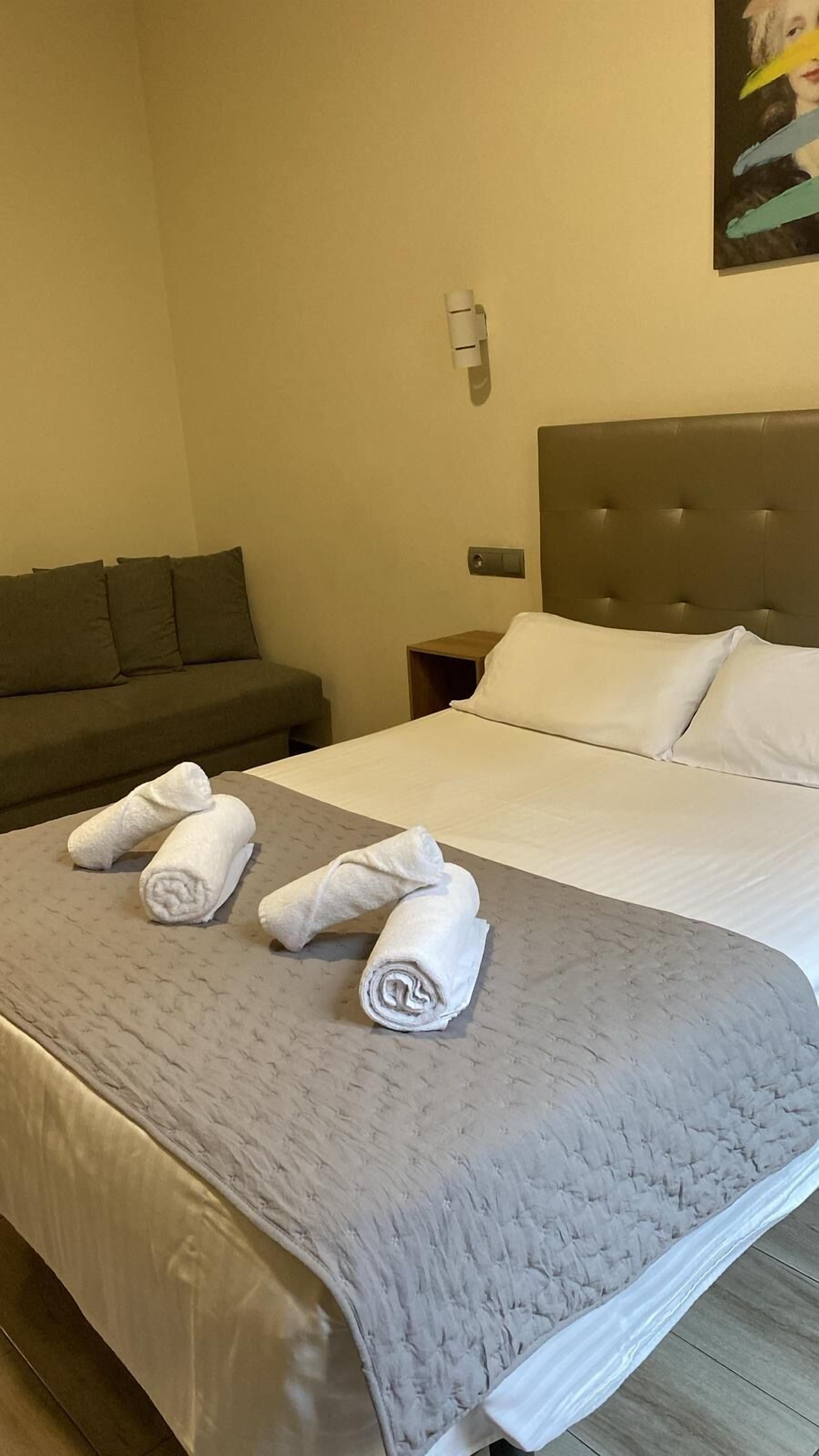 Habitación de hotel con una cama grande, una manta gris y tres toallas dobladas sobre la cama.