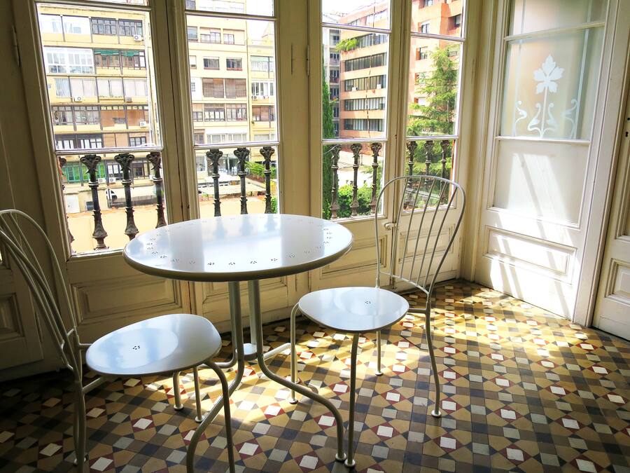 Una habitación con una mesa redonda y dos sillas, ventanas amplias con vistas a edificios y un suelo estampado.