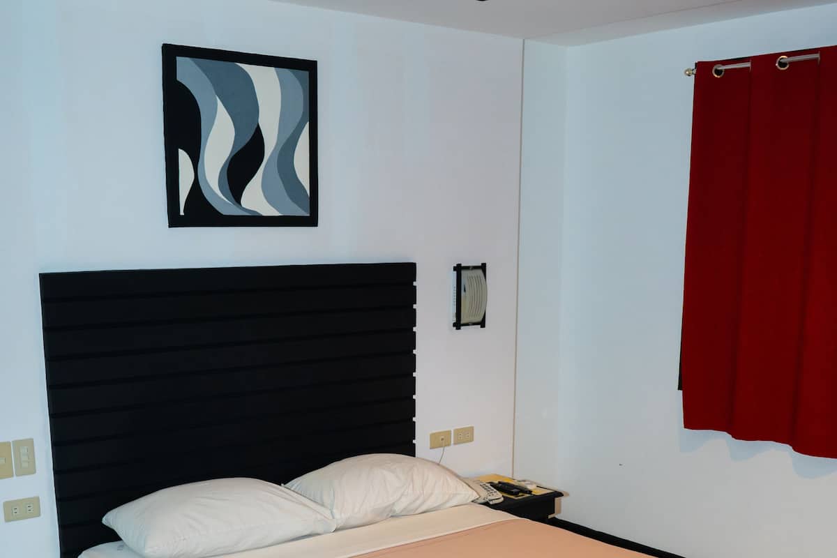 Un lit bien fait, avec une tête de lit noire, un rideau rouge et une œuvre d’art abstraite accrochée au mur.