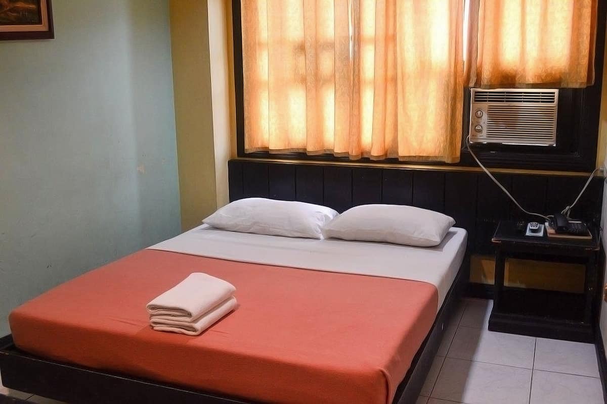Un lit simple avec un matelas rouge et des oreillers blancs, une petite table de chevet avec un téléphone et un chargeur, et une fenêtre avec des rideaux.
