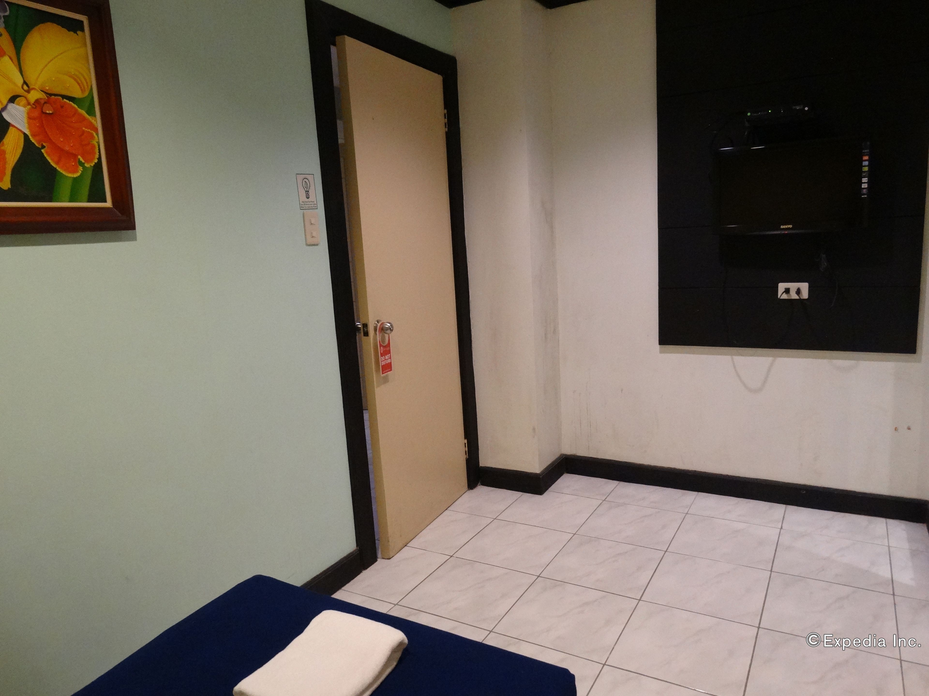 Een kamer met een blauwe massagetafel, een deur met een rode tag, een bloem in een lijst aan de muur, en een zwarte tv die aan de muur is bevestigd.