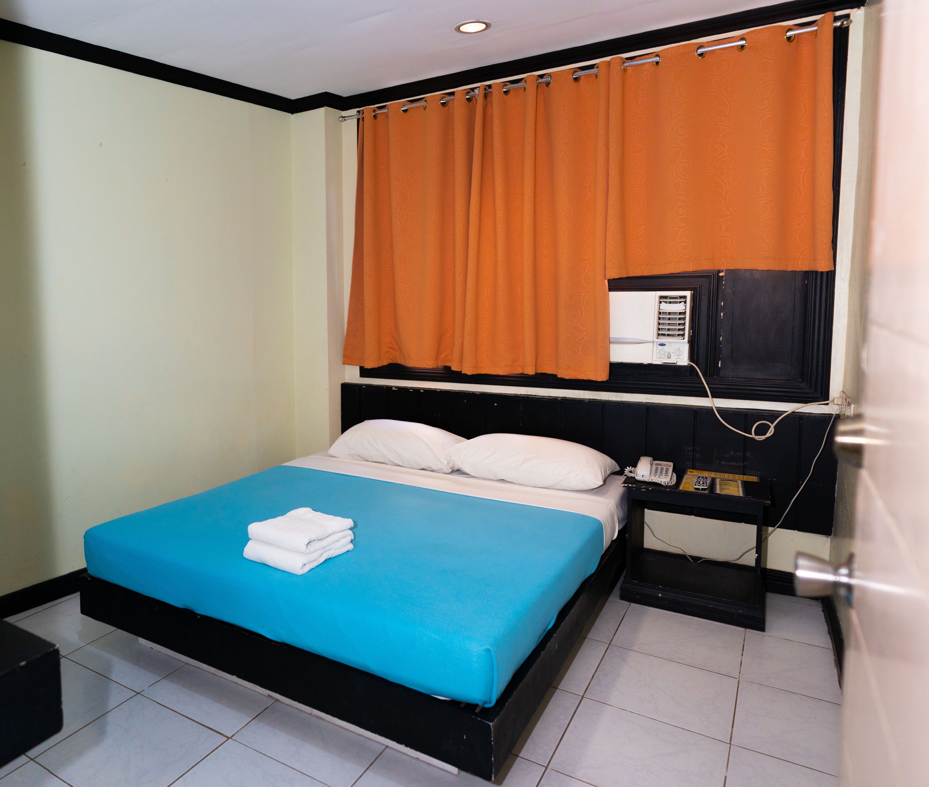 Una habitación con cama, mesita de noche, unidad de aire acondicionado y ventana con cortinas naranjas.