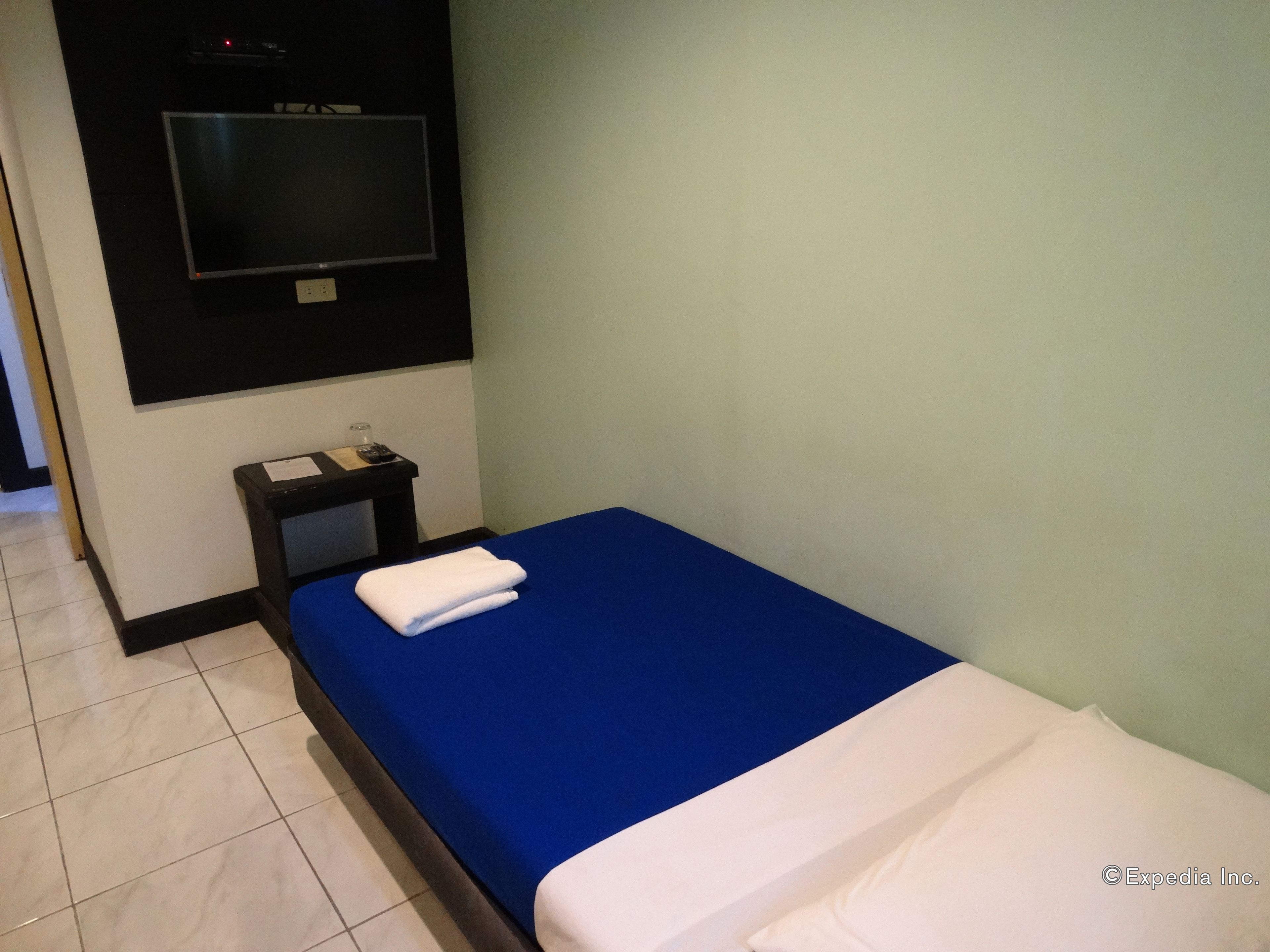A small, single-bed room with a blue bedspread, a white pillow, and a white towel folded on the bed. There is a small table with a lamp and a remote control on it. The room has light green walls and a black TV mounted on the wall.