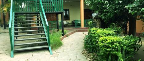 A courtyard with a stone staircase, a green metal railing, and lush greenery.