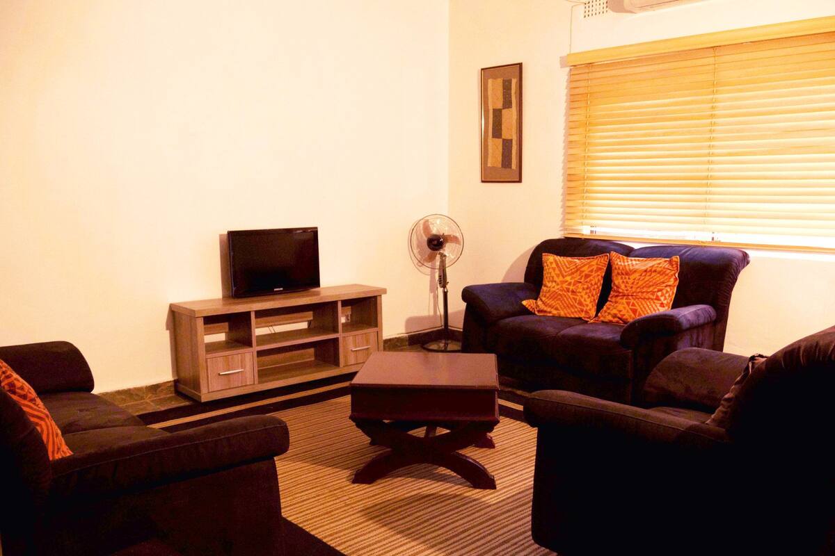 A living room with a TV on a wooden stand, a floor fan, and two sofas with patterned cushions.