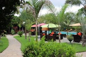 A poolside area with lounge chairs and umbrellas, surrounded by lush greenery and a stone pathway. - Palmwood Lodge (Lusaka)
