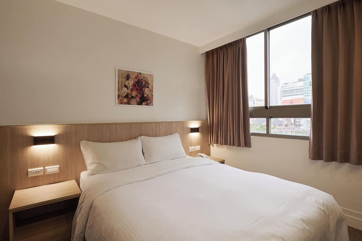 Quarto de hotel com uma cama grande, mesa de cabeceira, telefone fixado na parede e uma janela com cortinas.